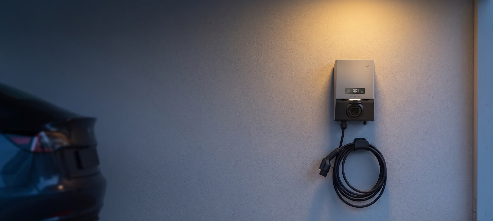 Wallbox an der Wand eines Carports mit aufgerolltem Ladekabel, daneben ein Elektroauto im Abendlicht
